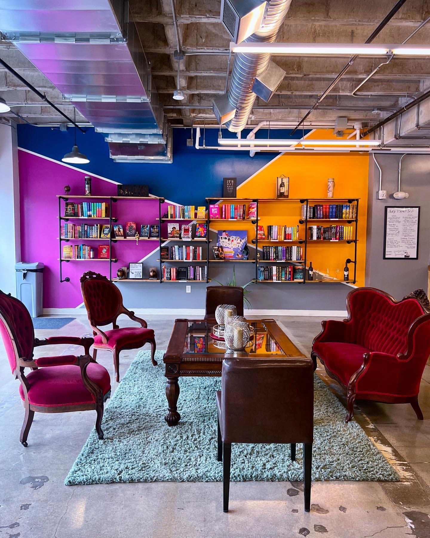 A vibrant room with colorful bookshelves filled with books against a purple and orange wall. Elegant red and brown chairs surround a wooden coffee table on a light green rug. Exposed ceiling with industrial decor.