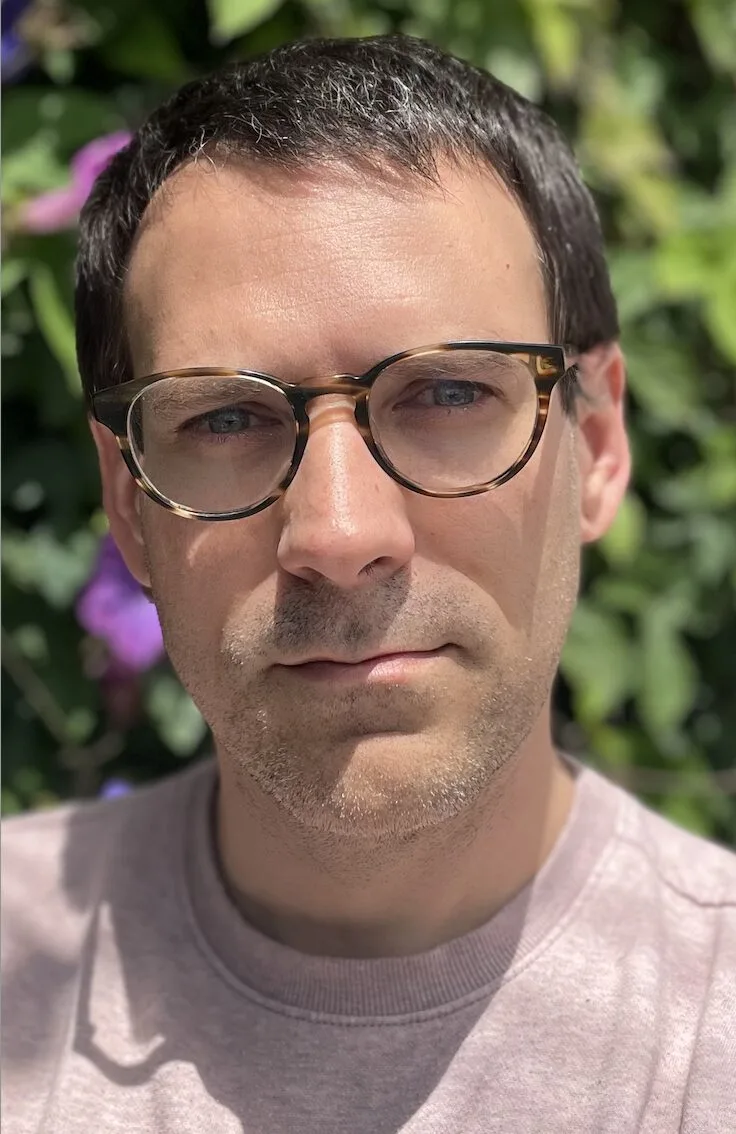 A person with short dark hair and glasses is looking at the camera. They are wearing a light-colored shirt. The background is filled with greenery and some purple flowers, suggesting an outdoor setting.