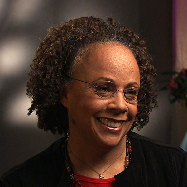 Person with curly hair and glasses, smiling and wearing a dark top with a beaded necklace. The background is softly lit with abstract patterns and a hint of foliage on the side.