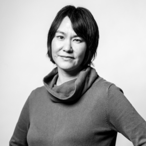 Woman with short dark hair wearing a high-collared sweater, standing with one hand on her hip, smiling softly, in a black and white portrait.