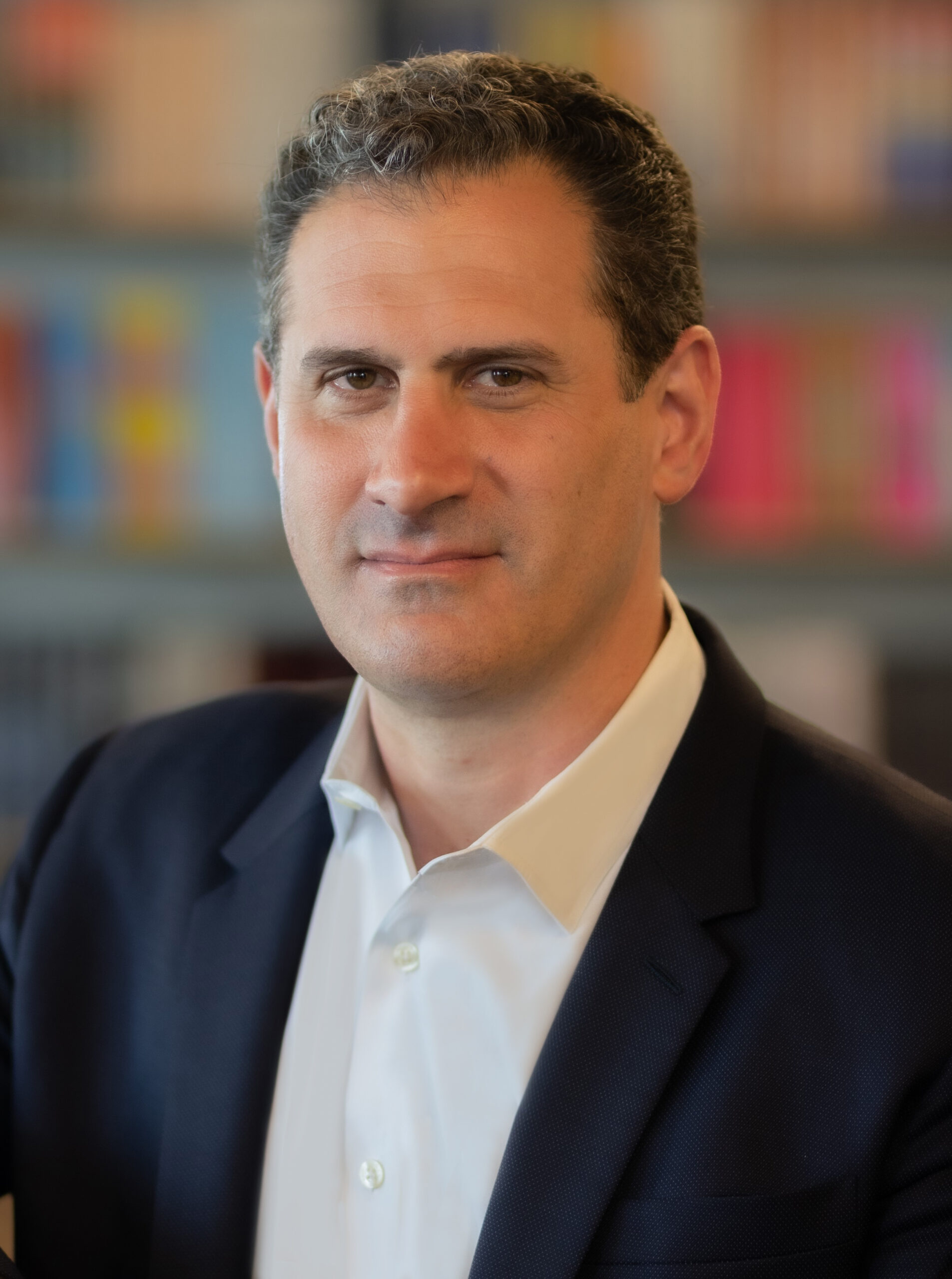 A person with short, curly hair, wearing a white shirt and a dark blazer, is standing in front of a blurred background of bookshelves. The expression is neutral and the setting appears to be a library or study.
