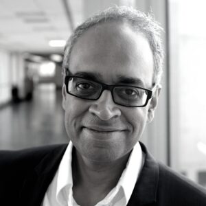 A person wearing glasses and a suit jacket smiles while standing in a corridor. The background is blurred, and the image is in black and white.