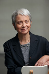 An older adult with short gray hair, known among world literature speakers, wears a dark blazer over a gray top. They smile slightly, resting their arm on the back of a white chair against a neutral background.