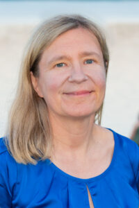 A woman with shoulder-length blonde hair, a renowned speaker in world literature, smiles softly while wearing a blue top. The background is blurred, with a light color that might be a beach or sky.