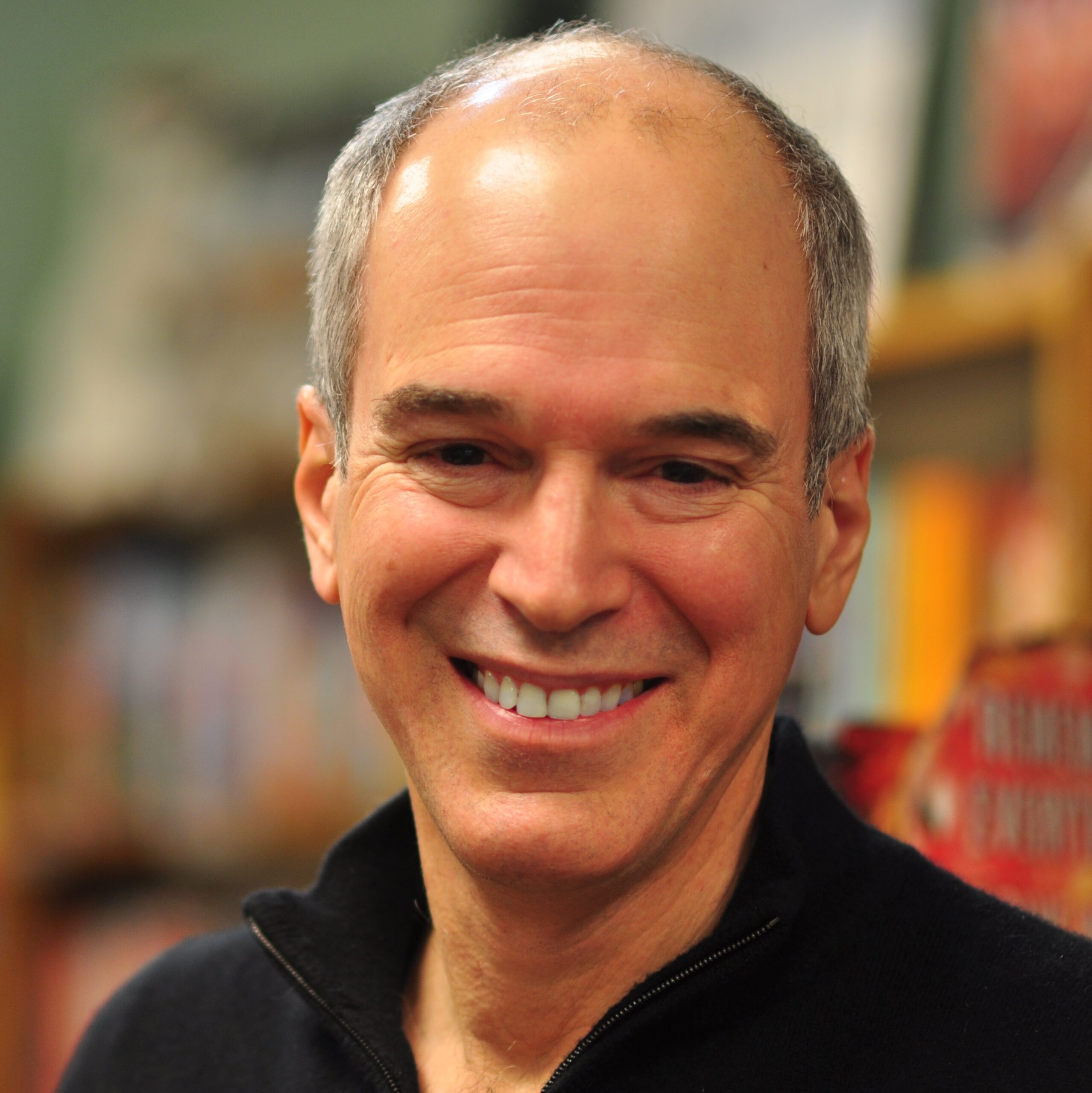A smiling man with short gray hair wearing a dark zip-up sweater. He stands in a room with bookshelves in the background.