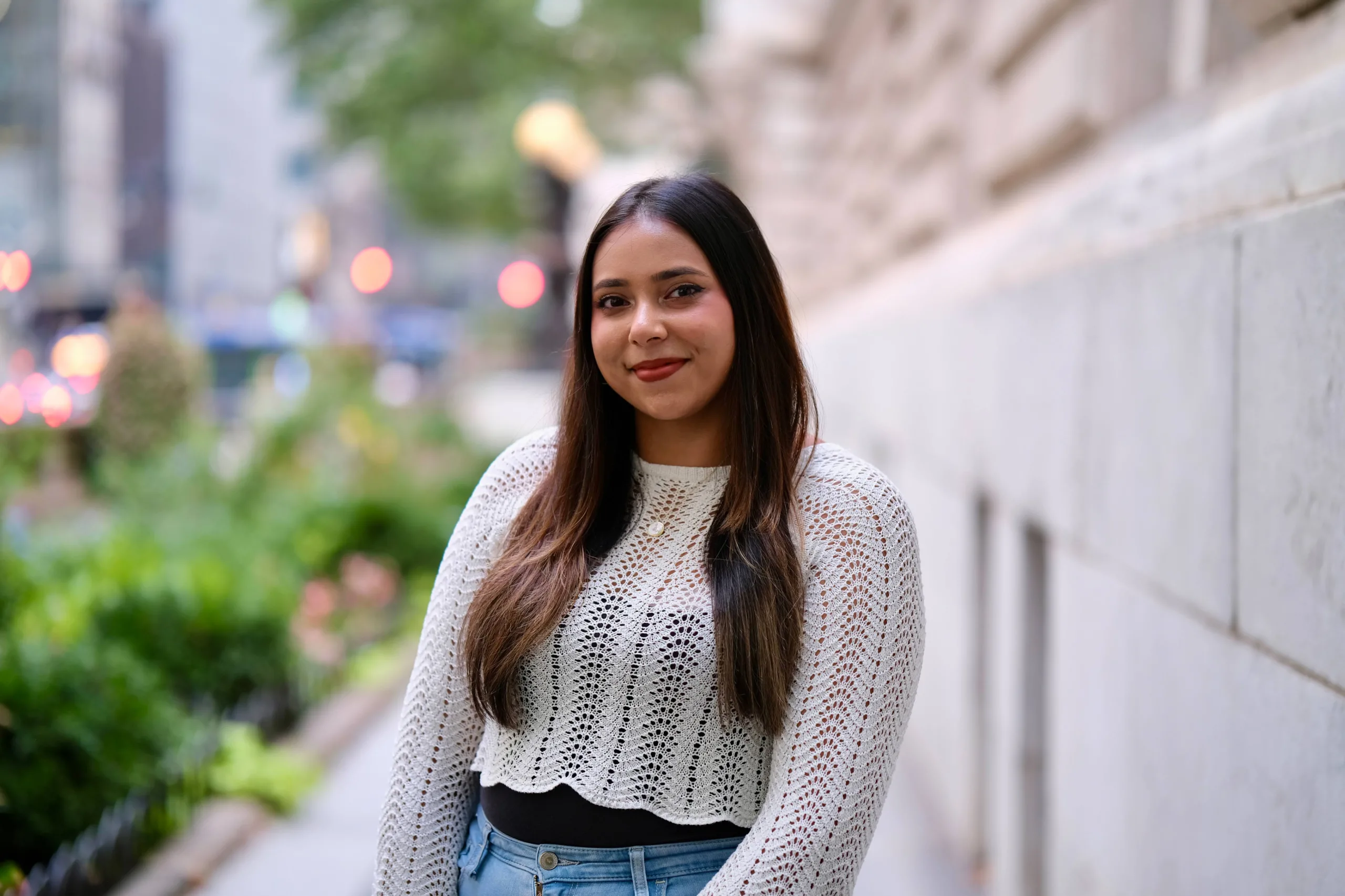 A woman with long dark hair stands outdoors, wearing a white knit top and jeans. She is smiling with a blurred cityscape and greenery in the background.