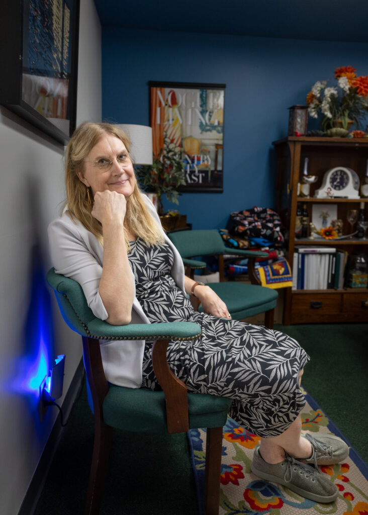 A woman in a patterned dress and light cardigan sits on a green chair in a cozy room, reminiscent of Jennifer Finney Boylans chic style. Her hand rests under her chin amid blue walls, eclectic decorations, and colorful accents. A cabinet and framed artwork complete the scene.