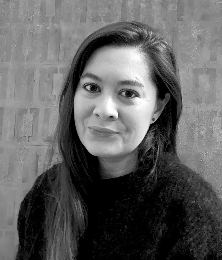 Black and white portrait of a woman with long hair, wearing a dark, fuzzy sweater. She is looking at the camera with a slight smile. The background features a textured wall.