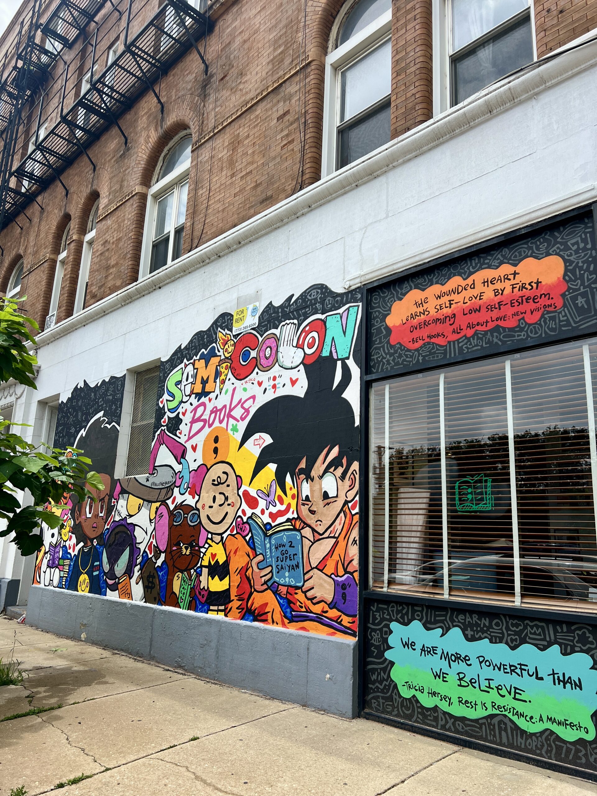 Colorful mural on a bookstore wall featuring animated characters and the name Semicolon Books. The store windows display a motivational quote about strength and healing. The building has a brick facade and an exterior staircase.