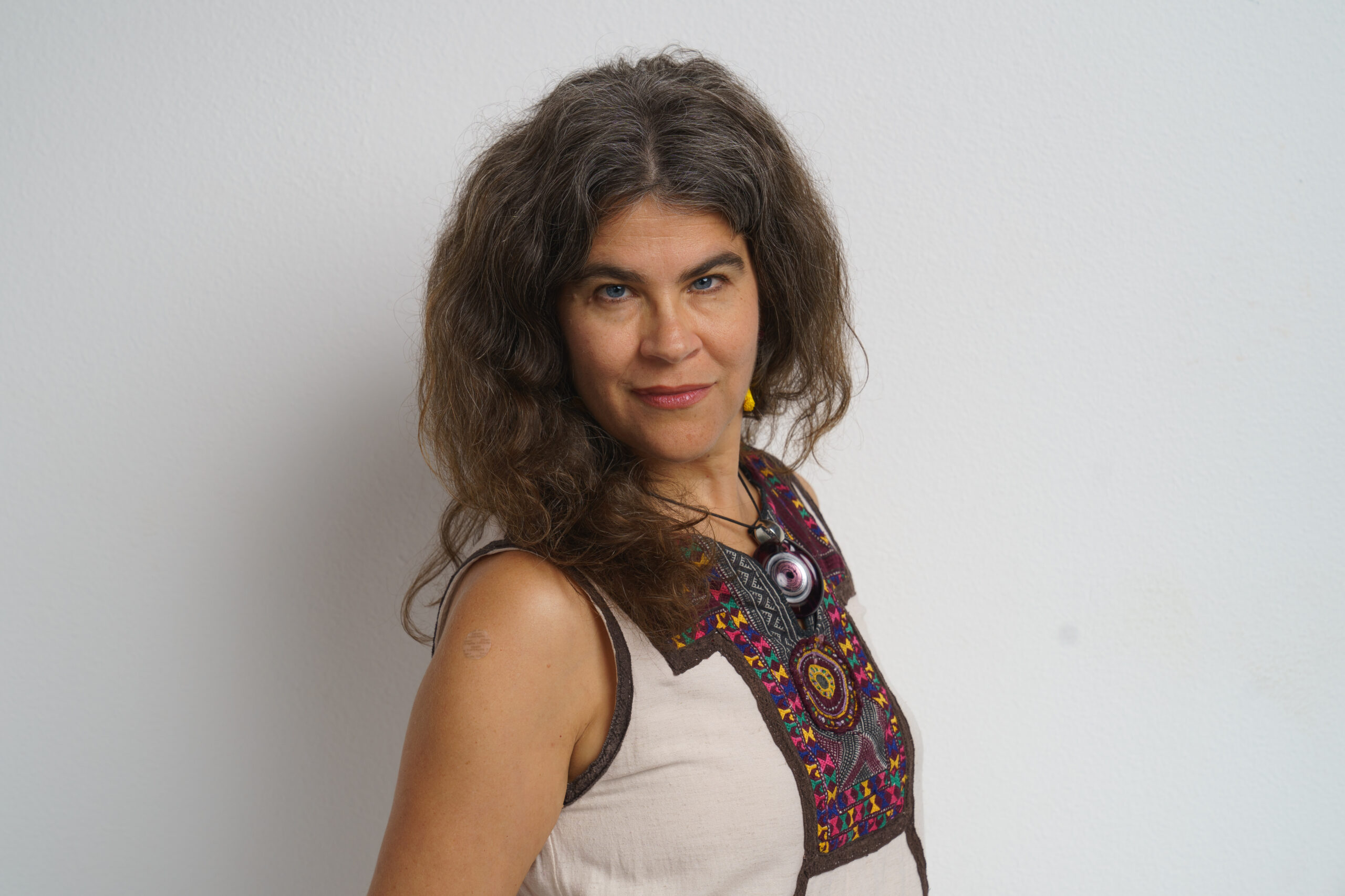 A woman with long, wavy hair stands against a white background. She wears a sleeveless top with colorful embroidery and a large pendant necklace. She is looking directly at the camera with a slight smile.