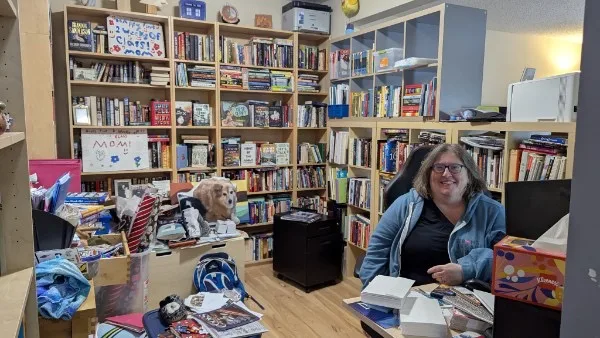 A person sits at a desk in a room filled with bookshelves. The shelves are packed with various books and games. A sign with Mom! is visible, along with a poster of a dog. The desk has assorted papers and items.