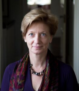 Portrait of a person with short, light brown hair, wearing a dark purple top and a colorful scarf. The softly blurred background, reminiscent of an indoor gathering for world literature speakers, frames their direct gaze and neutral expression perfectly.