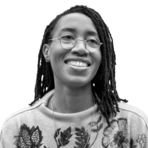 A person with long dreadlocks, wearing glasses and a sweater with floral patterns, smiles while looking slightly upwards. The image is in black and white.