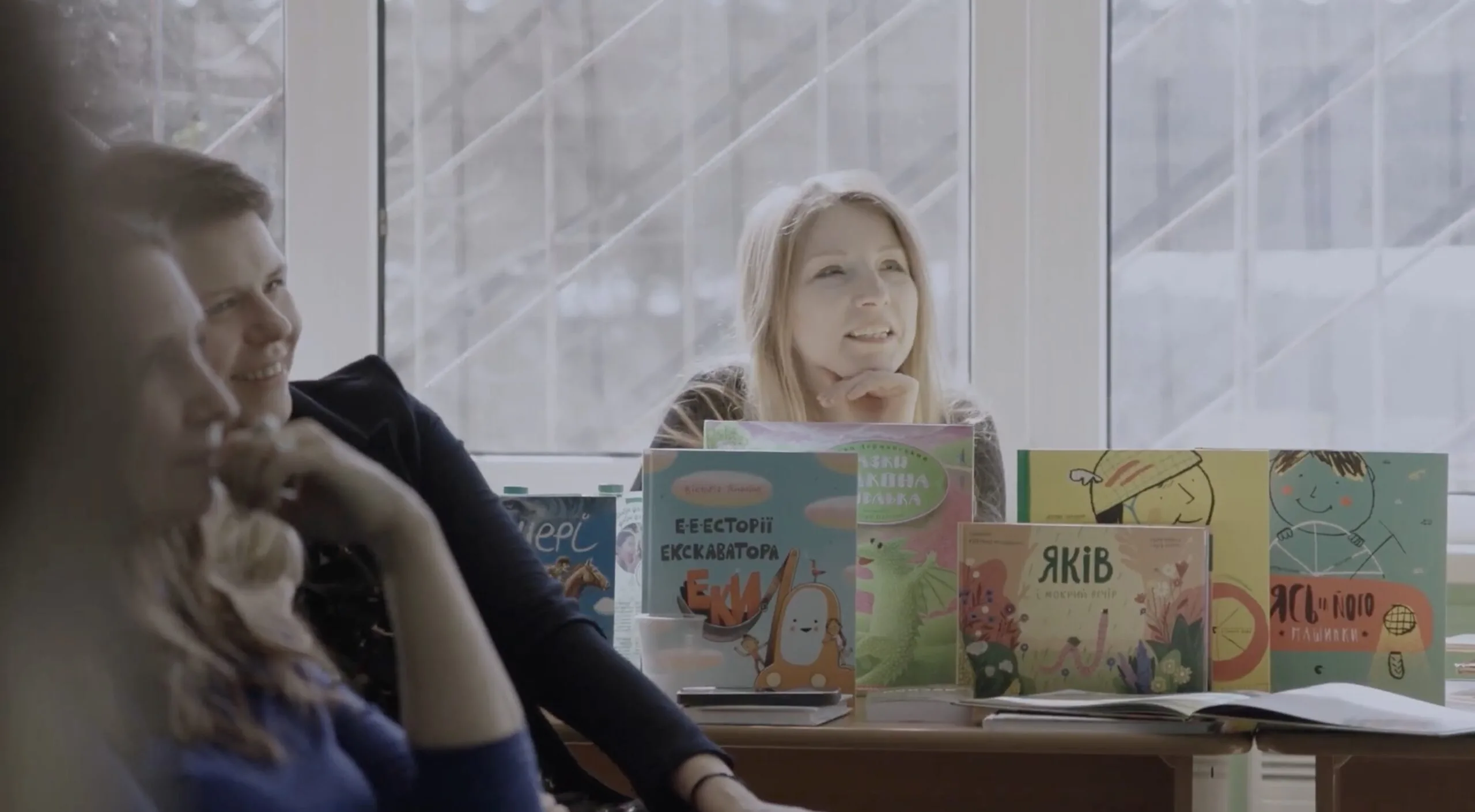 Three people sit at a table filled with colorful childrens books. A woman in the center, smiling, looks attentively forward. Natural light from a large window brightens the room, creating a relaxed atmosphere.