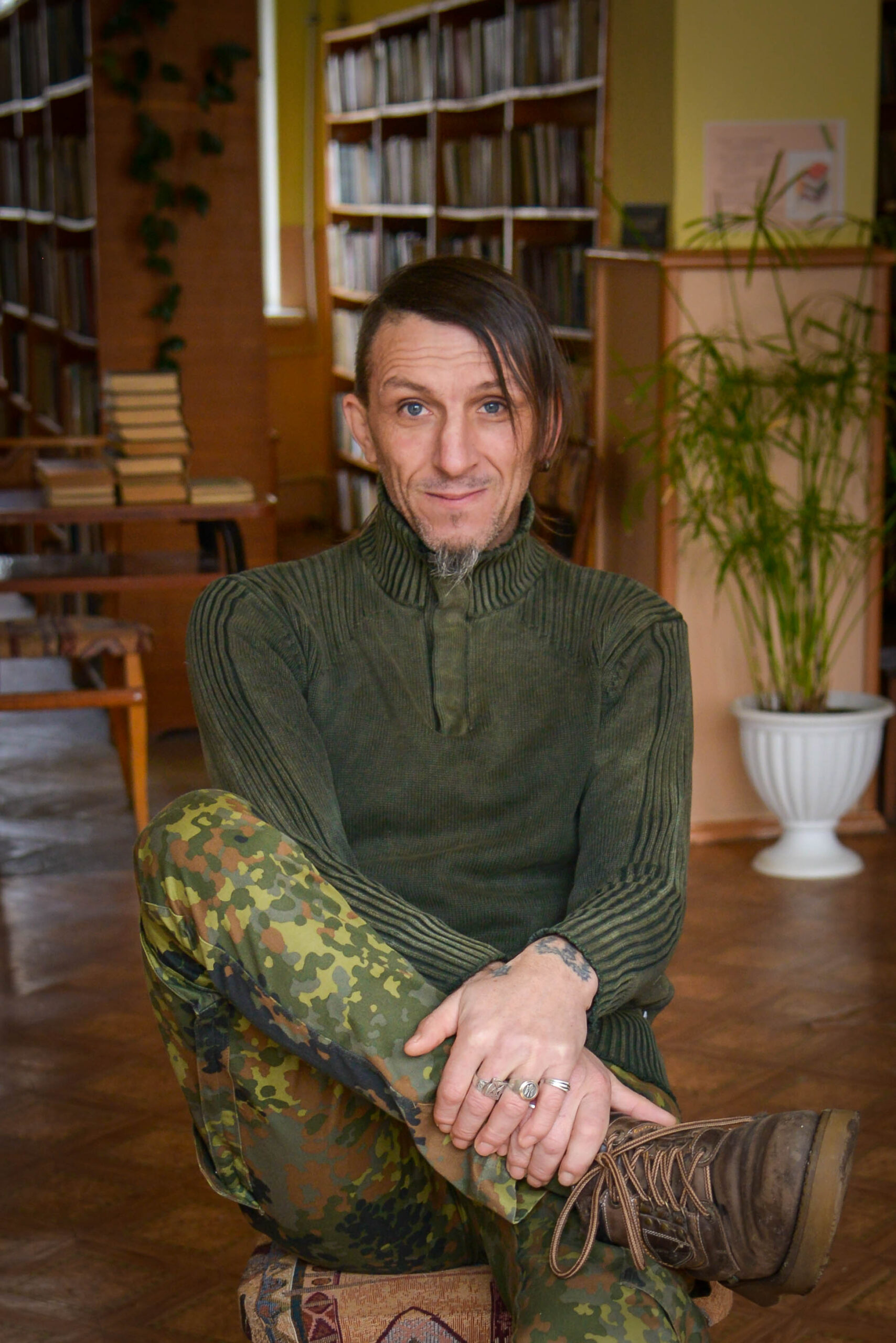A person with shoulder-length hair and stubble sits cross-legged on a wooden chair in a library. They wear a green sweater and camouflage pants, with brown boots, smiling at the camera. Bookshelves and a potted plant are in the background.