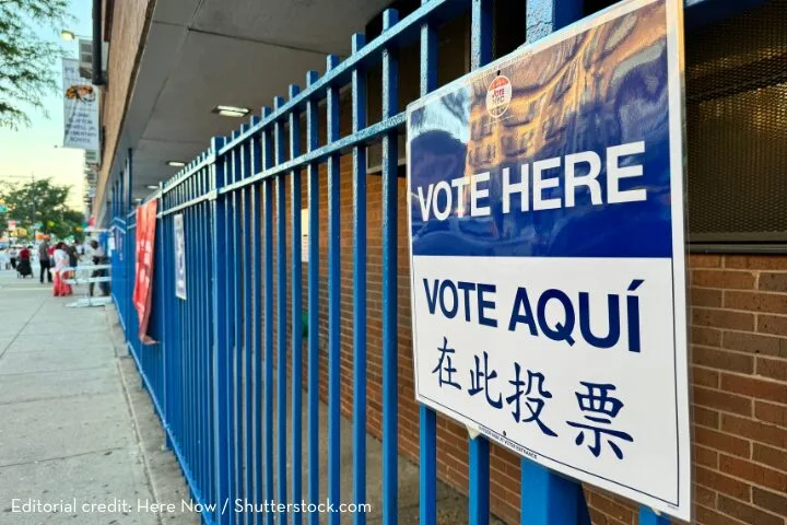 Blue metal fence with a multilingual Vote Here sign in English, Spanish, and Chinese. People in the background are entering a brick building, presumably a polling station, with another flag visible along the fence.