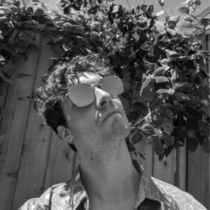 A black-and-white photo of a person with curly hair wearing reflective sunglasses, looking upwards. They are standing in front of a wooden fence covered with leafy vines.