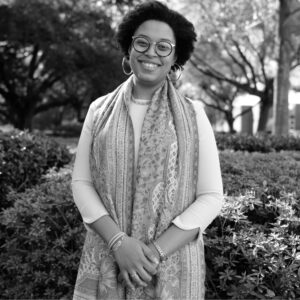 A smiling person stands outdoors in front of bushes and trees. They have curly hair, wear glasses, and are dressed in a light long-sleeve shirt with a patterned scarf. The image is in black and white.