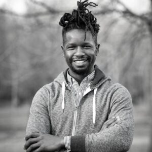 Smiling man with dreadlocks, wearing a hoodie over a checkered shirt, stands in an outdoor setting. Arms crossed. Black and white photo.