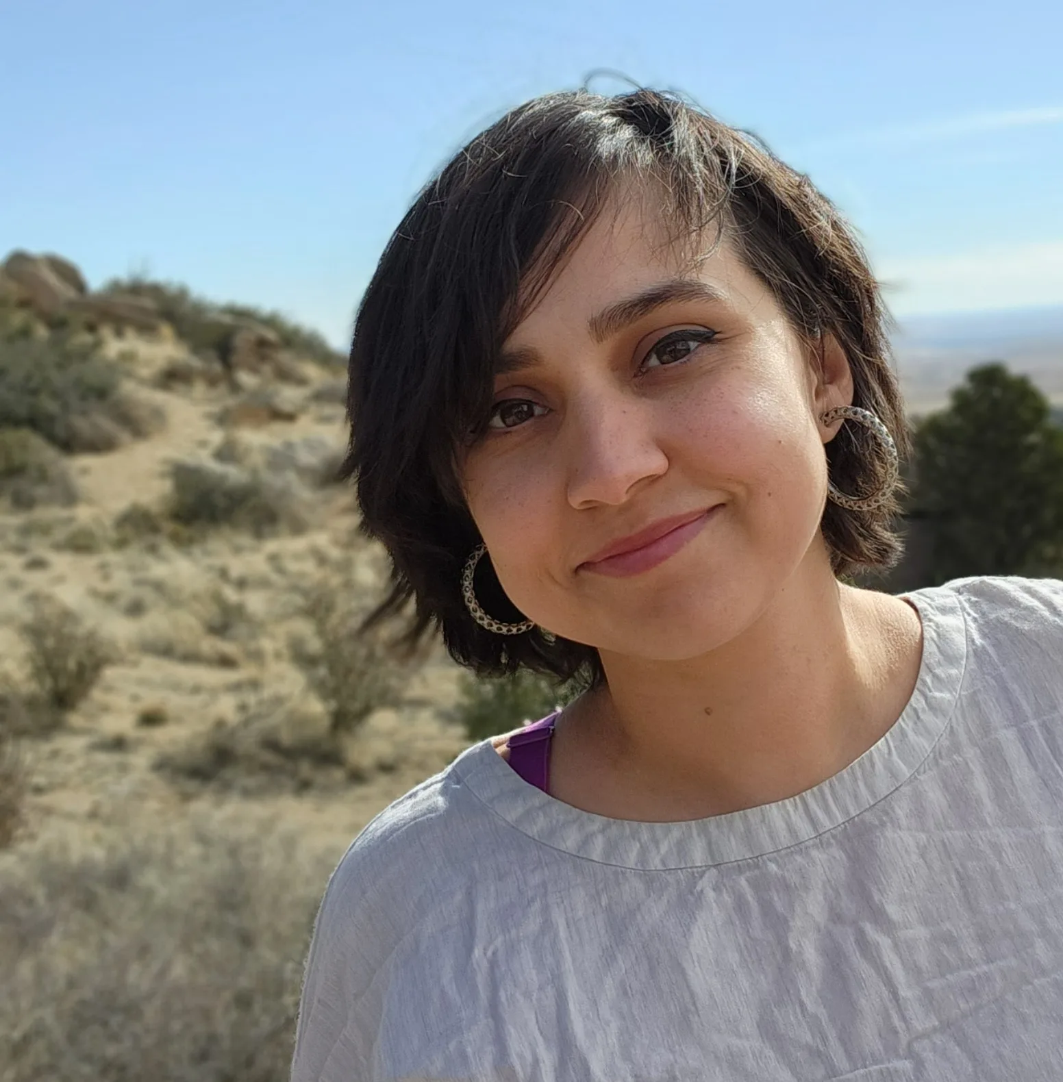 A person with short dark hair is smiling and wearing a light-colored top and hoop earrings. The background features a sunny outdoor landscape with dry shrubs and hills.