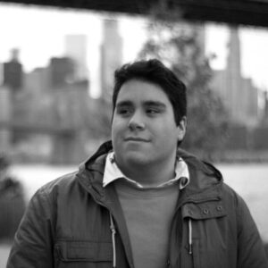 A person with short dark hair is standing outdoors, wearing a jacket and sweater. The background is blurred, showing a bridge and city skyline. The photo is in black and white.