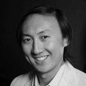 A black and white portrait of a smiling person with shoulder-length hair. They are wearing a light-colored blazer and a collared shirt, set against a dark background.