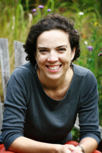 A beaming individual with short curly hair and a gray top sits outdoors in a garden, surrounded by tall green plants and purple flowers, like a scene from world literature.