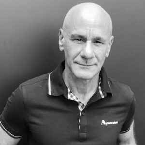 A bald man wearing a short-sleeved polo shirt with a checked collar stands against a plain dark background. The shirt has an A logo on the chest. He is looking at the camera. The image is in black and white.