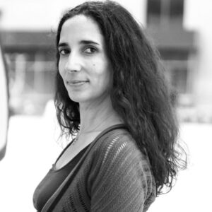 A black and white photo of a woman with long, curly hair, wearing a textured cardigan over a top. She is looking at the camera, standing in an urban setting with blurred buildings in the background.