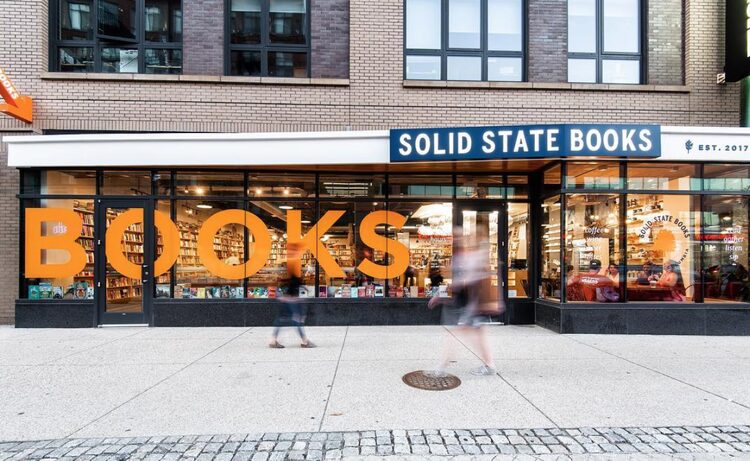 The image shows the exterior of Solid State Books, featuring large orange BOOKS signage on the window. Two people walk past the storefront on the sidewalk. Inside, shelves filled with books are visible through the glass.