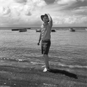 A person wearing a wide-brimmed hat and casual clothing stands barefoot on a beach with gentle waves. They hold the hat with one hand, looking towards the ocean. Several boats float in the distance under a partly cloudy sky. The image is in black and white.