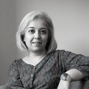 A person with short, light-colored hair sits in a chair, smiling. They are wearing a patterned top and a watch. The background is a plain wall, and the image is in black and white.