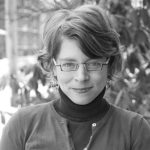 A black and white portrait of a person with short hair, wearing glasses, a turtleneck, and a sweater. The person is outdoors with foliage in the background. They have a slight smile and are facing the camera.