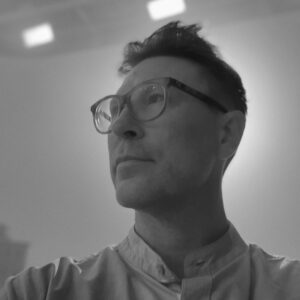 Black and white photo of a person wearing glasses and a collared shirt, looking to the left. Soft lighting creates a halo effect around their head, with a minimalist background.