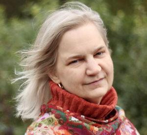 A person with long, light hair and a thoughtful expression stands outdoors, resembling a speaker from world literature. They are wearing a colorful scarf and a red turtleneck sweater against the backdrop of blurred greenery.
