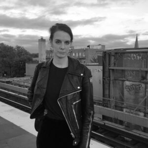Black and white photo of a person wearing a leather jacket standing on a platform with urban buildings and graffiti in the background. The sky is cloudy, and the overall mood is moody and contemplative.