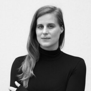 A person with long hair wearing a black turtleneck stands against a plain light background, looking directly at the camera with a neutral expression.