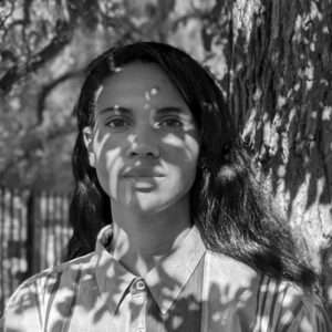A person with long hair stands against a tree trunk. Dappled shadows of leaves are cast across their face and shirt. The background features blurred foliage and a fence. The image is in black and white.