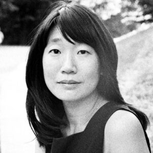 Black and white portrait of a person with long dark hair and a serene expression, wearing a sleeveless top. They are outdoors with a blurred background of grass and trees.