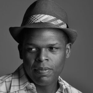 A black and white portrait of a person wearing a plaid shirt and a fedora with a striped band. The person is looking off to the side with a slight smile, set against a plain background.