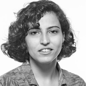 Black and white portrait of a person with short, curly hair and a slight smile. They are wearing a patterned shirt and looking directly at the camera against a plain background.