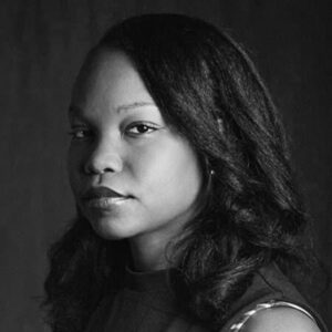 Black and white portrait of a person with long, dark hair, gazing directly at the camera. The background is plain and dark, emphasizing the subjects calm and composed expression.