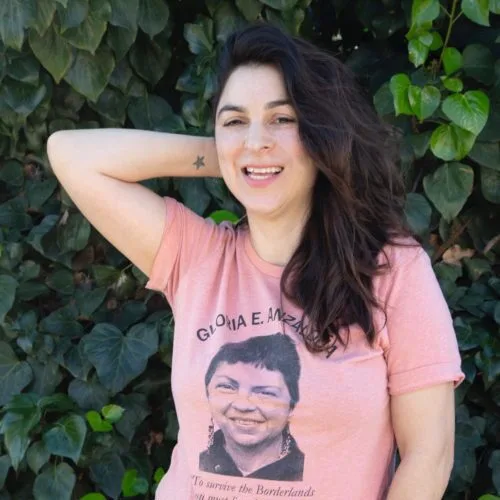 Woman smiling with one hand behind her head, wearing a pink T-shirt featuring a printed image of Gloria E. Anzaldúa. The background is lush with green leaves.