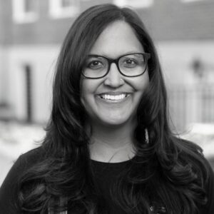 A person with long, dark hair and glasses smiles warmly at the camera. They are wearing a dark top. The background is blurred and appears to show a building and some outdoor space. The image is in black and white.