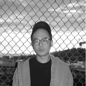 A person wearing glasses and a cap stands in front of a chain-link fence. The background shows a cloudy sky with hills. The person looks directly at the camera with a neutral expression. The image is in black and white.