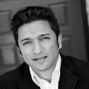 Black and white portrait of a person wearing a dark blazer and a white shirt, looking at the camera with a neutral expression. A closed door is in the blurred background.