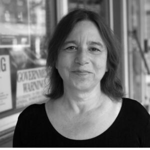 A woman with shoulder-length hair smiles softly, standing outdoors in front of a building. She wears a black top, and there are blurred signs and windows in the background. The image is in black and white.