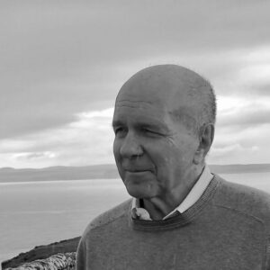 Black and white photo of an older man with a bald head, wearing a sweater, looking into the distance. The background features a serene seascape with a cloudy sky and distant hills.