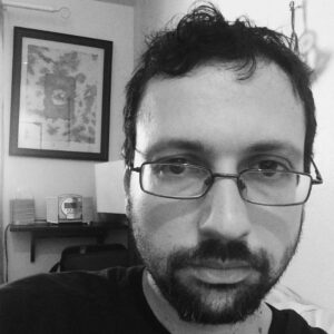 A black and white close-up photo of a person with glasses and a beard looking at the camera. In the background, theres a framed picture on the wall and a small shelf with books and other items.
