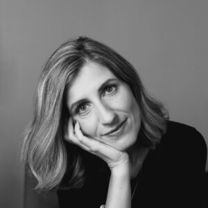 Black and white portrait of a person with shoulder-length hair resting their head on their hand, looking at the camera with a gentle expression. Theyre wearing a dark top and theres a neutral background.
